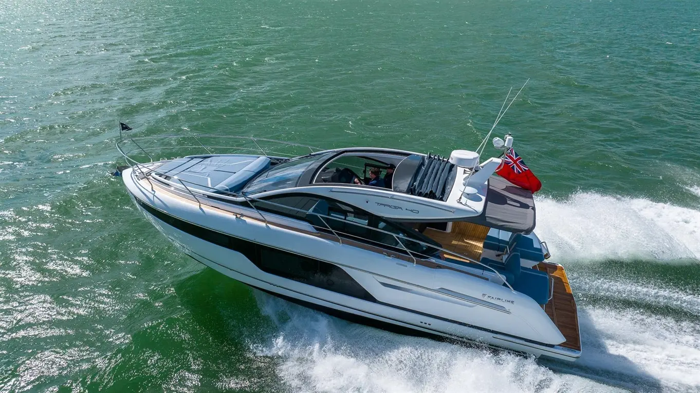 A sleek Fairline Targa 40 yacht glides through turquoise waters, leaving a white wake; a British flag flies from the stern.